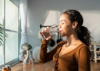 Young beautiful Latino woman holding clean water into glass in kitchen. Attractive active thirsty girl drink or take a sips of mineral natural in cup for health care and wellbeing in kitchen in house.
